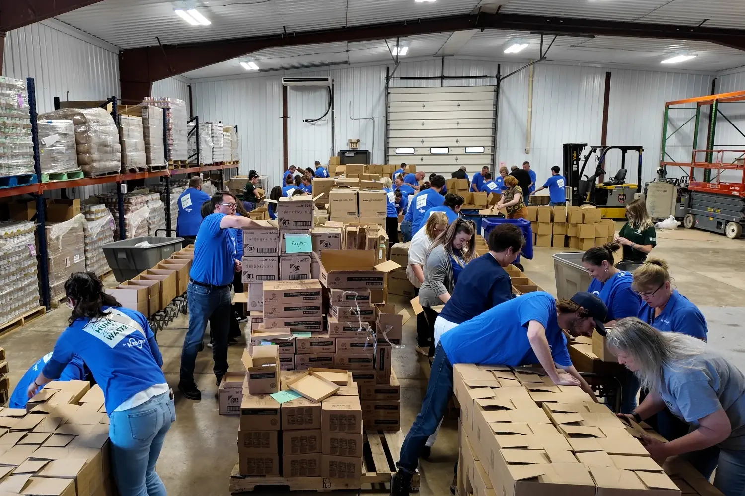 Dozens of Kroger team members in blue shirts working together in a warehouse, assembling food boxes from stacks of supplies and filling them along long rows of tables and conveyors.