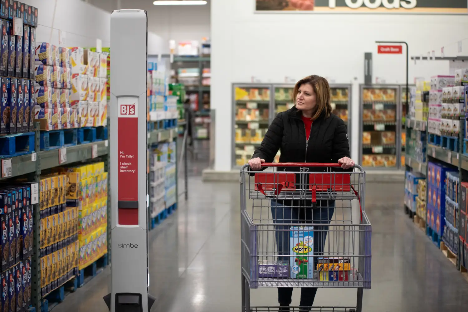 A woman pushes her cart through a grocery lane while a BJs's inventory robot scans product on the shelves.