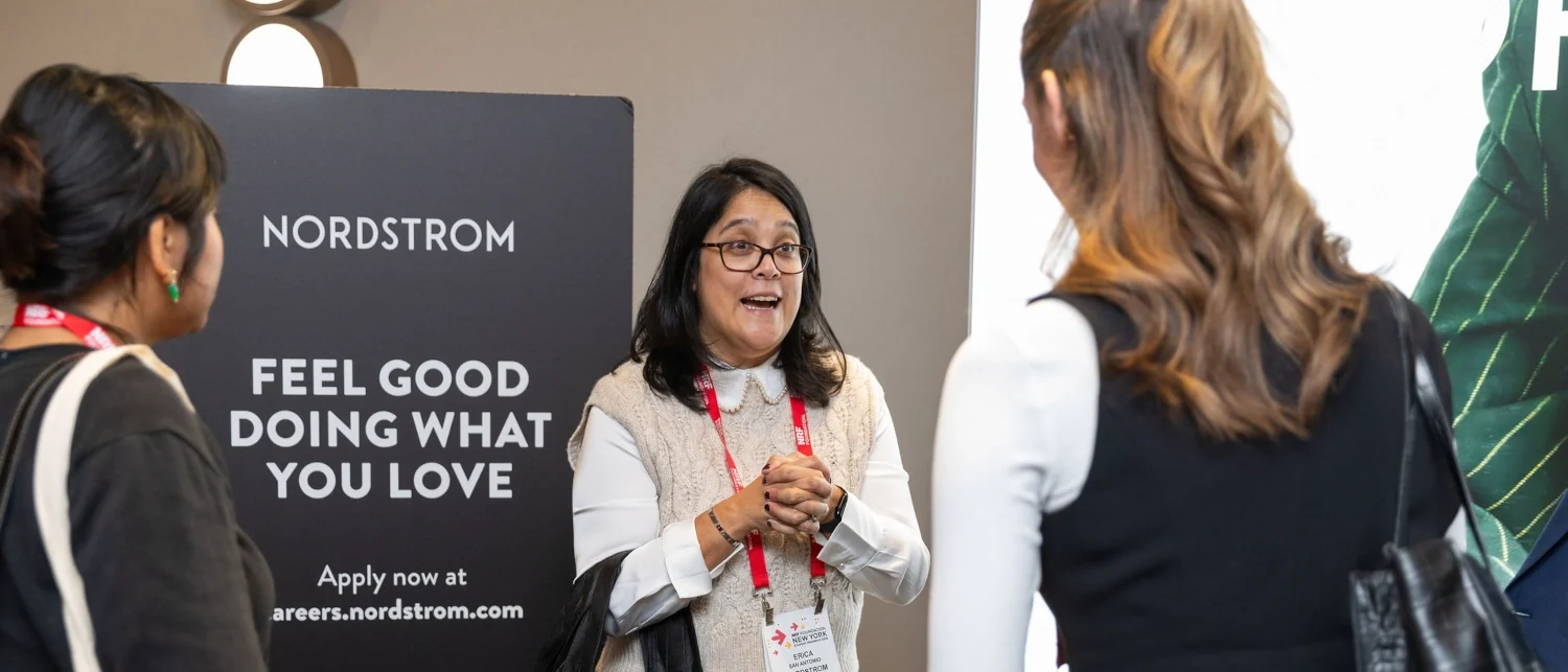 A Nordstrom recruiter talks to interested students at the NRF Foundation Student Program Career Fair. The recruiter wears a laynard around her neck in front of a sigh that says Nordstrom: Feel Good Doing What You Love.