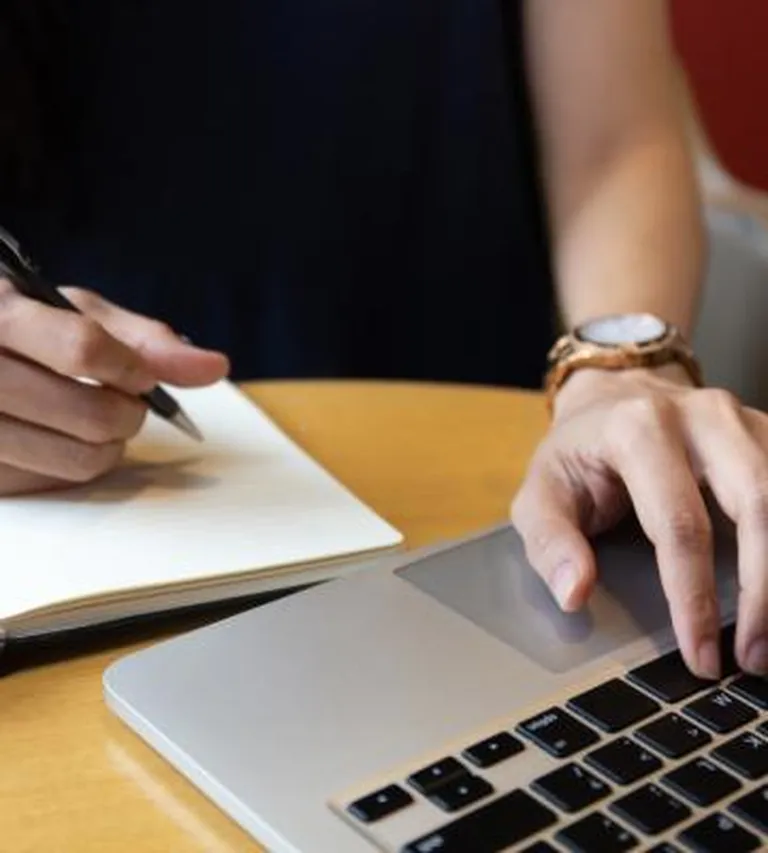 Someone typing on a laptop and taking notes in a journal.