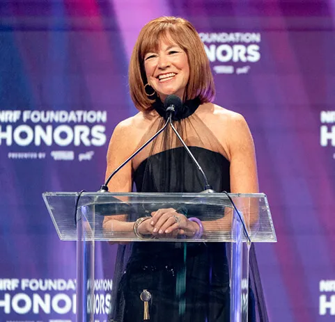 Barbra Bukovac in sheik evening wear speaks on stage at the Honors '26 podium.