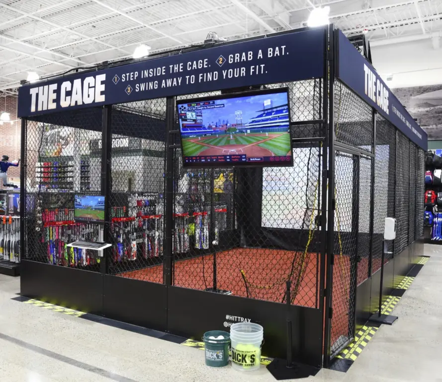 A batting cage is setup inside a DICK'S Sporting Goods store. The cage is enclosed with black netting and has a sign on top that reads 'THE CAGE' along with instructions: 'Step inside the cage. Grab a bat. Swing away to find your fit.'