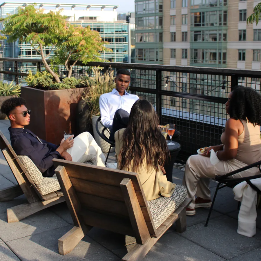 Four professionals sip cocktails and discuss the day's learnings at a rooftop bar. The setting sun is shining and casting shadows across tabletops.