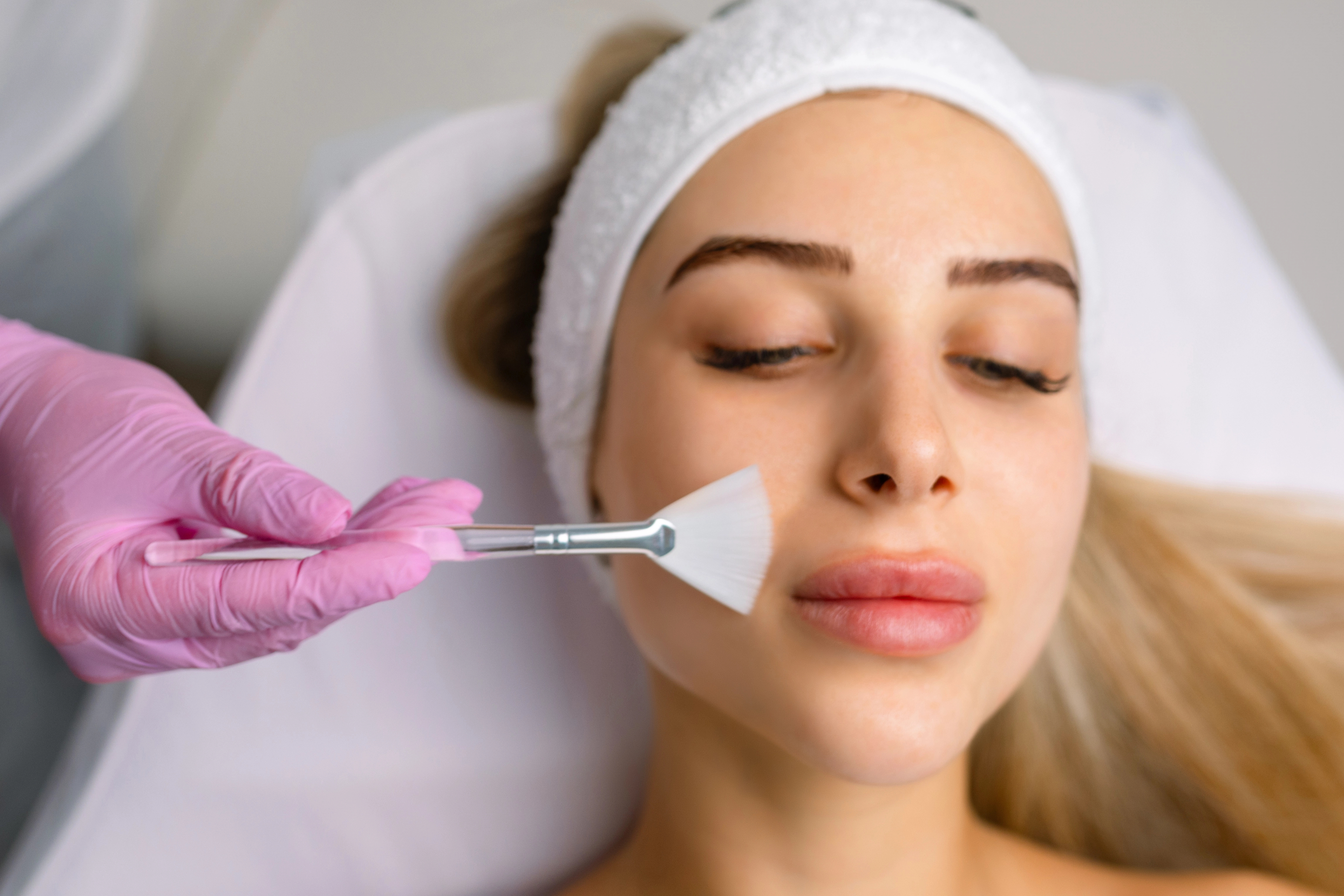 A woman receives a facial treatment, with a hand in pink gloves applying a mask using a brush. She is lying down with a white headband.