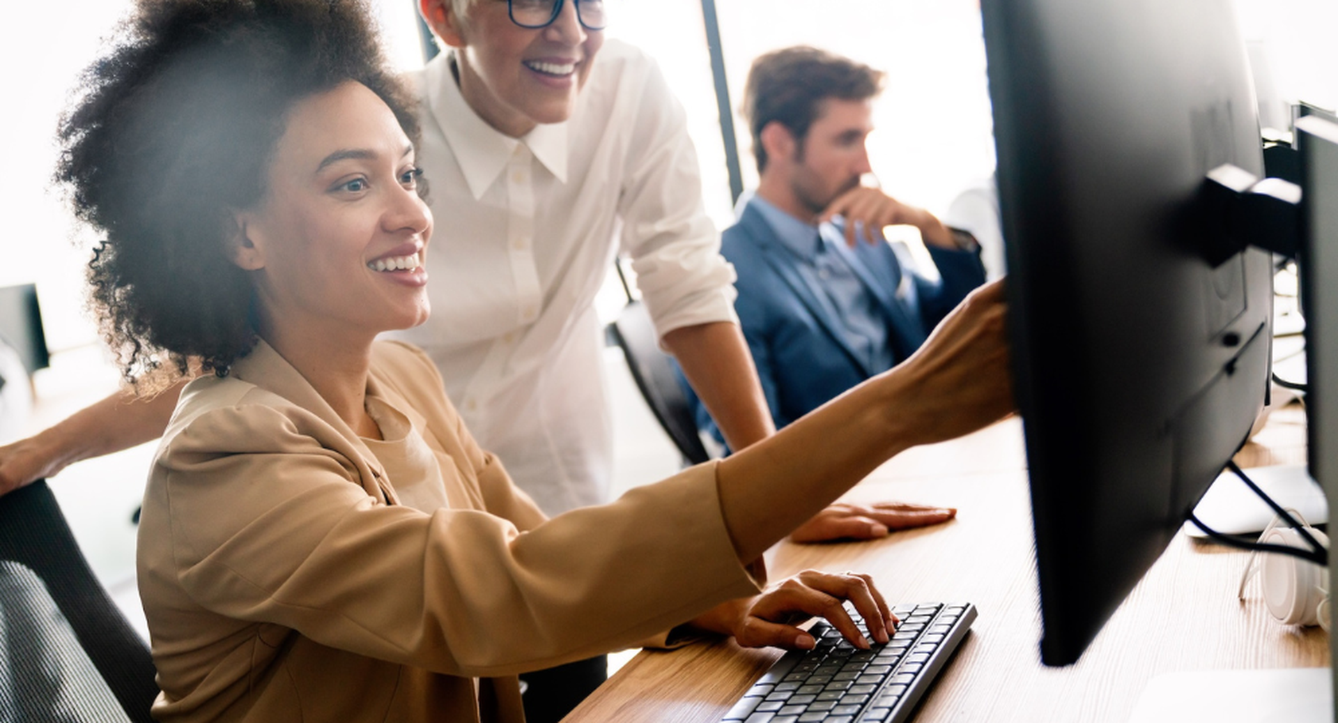 Two women smiling and collaborating at a computer in an office, while a man sits focused in the background.