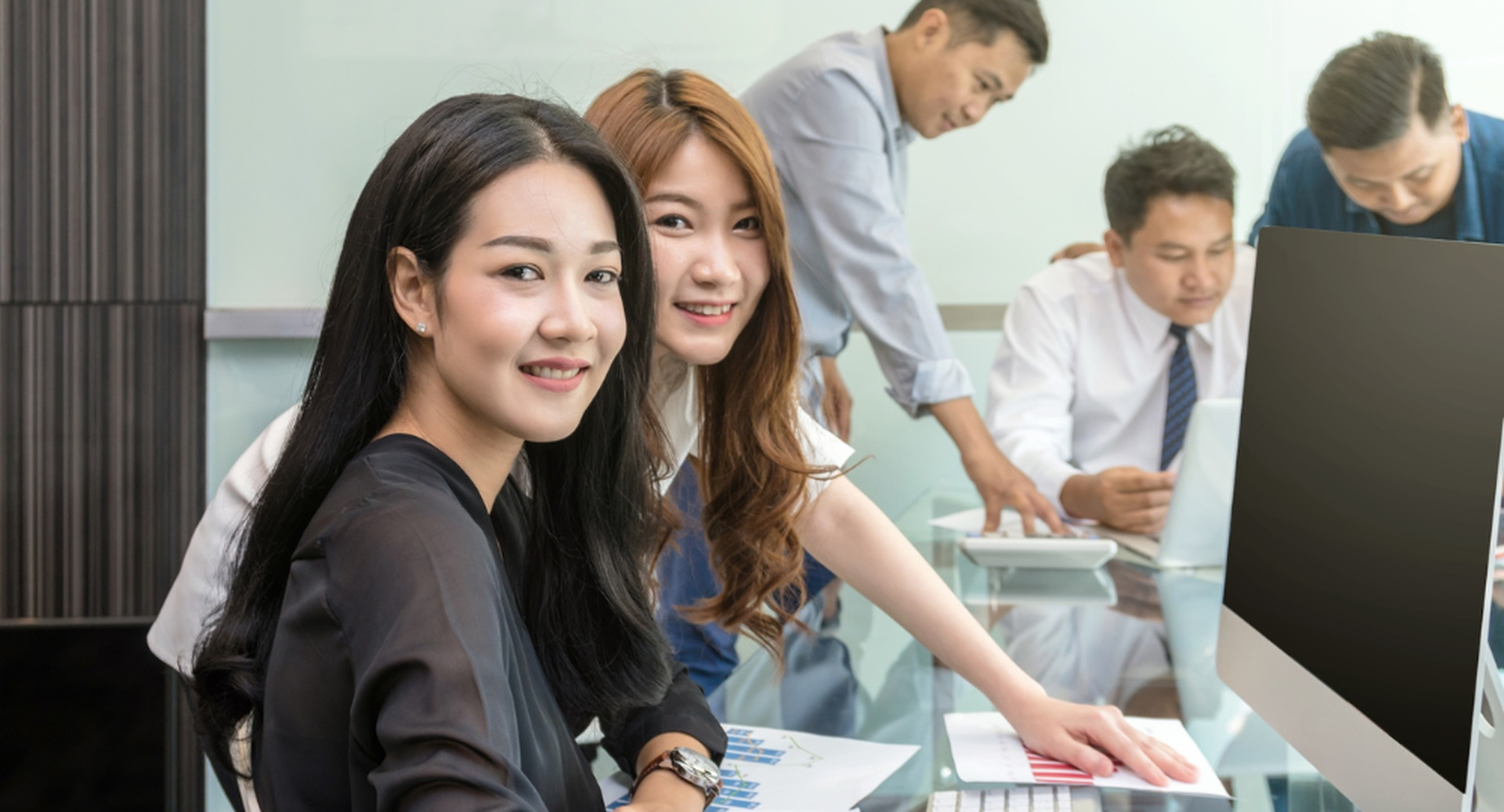 Two women smile at the camera, seated in front of a computer, while three men work in the background in an office setting.