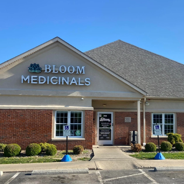 Bloom Medicinals storefront with lotus logo on beige brick building, central glass door, walkway and two handicap parking signs under clear blue sky.