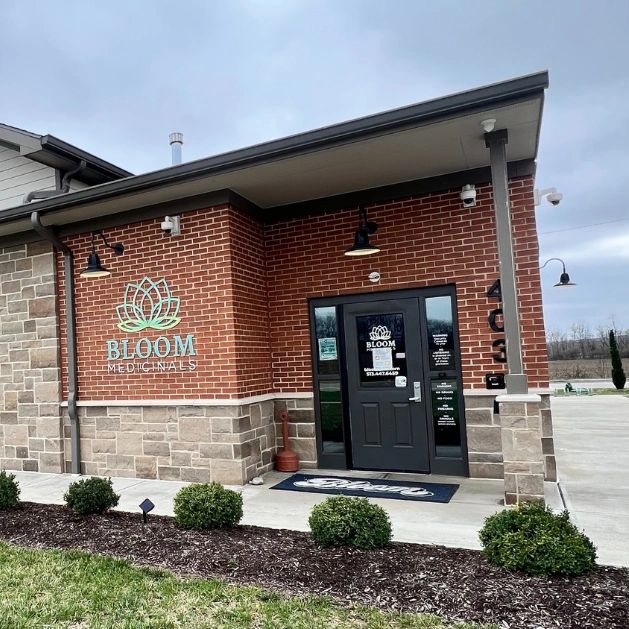 Exterior of Bloom Medicinals storefront on brick building with lotus logo, glass door, and small landscaped shrubs.