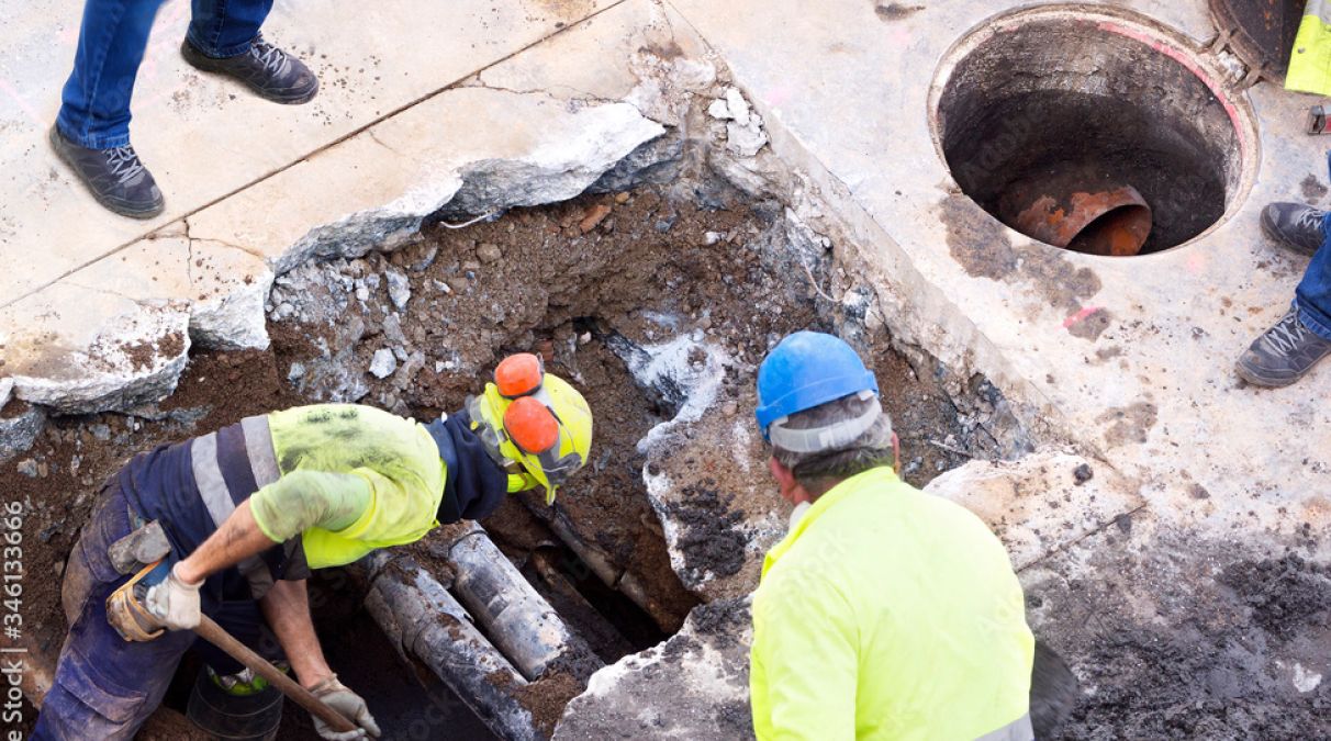 Several construction workers wearing safety helmets and vests working in a deep pit near exposed pipes on a construction site.