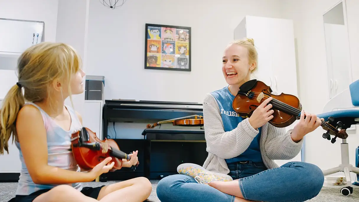 Musicologie teacher and student sitting cross-legged on the floor with violins, laughing.
