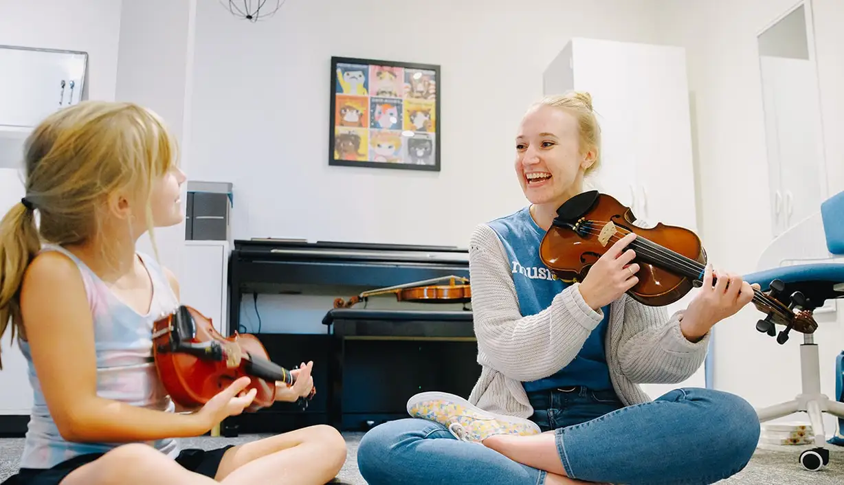 Musicologie teacher and student sitting cross-legged on the floor with violins, laughing.