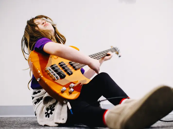 Child sitting on the floor with an electric bass guitar, leaning back in a playful pose.