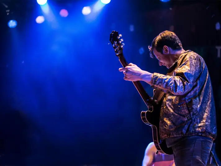 A man wearing a gold jacket while playing guitar under stage lighting