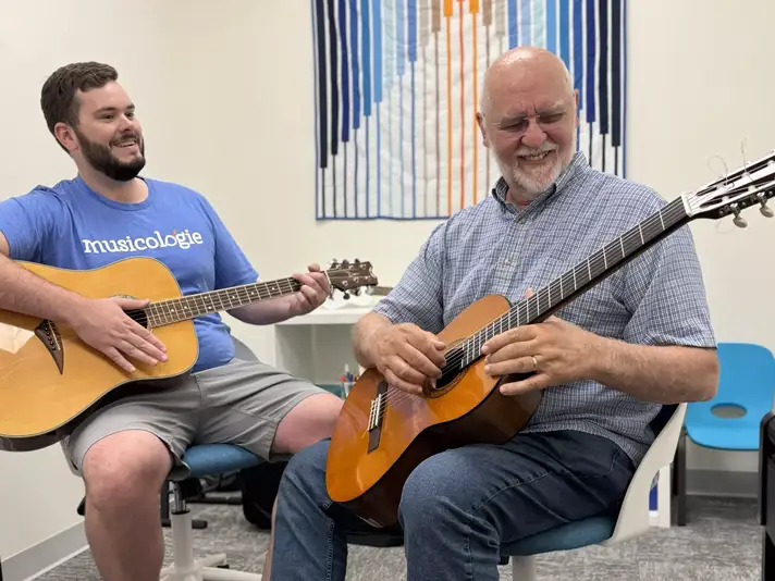 Older adult student smiling while playing acoustic guitar alongside a Musicologie teacher during a guitar lesson.
