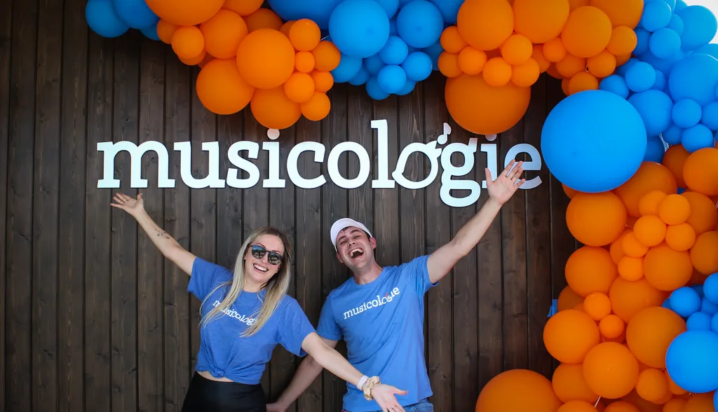 Founder Kay Barker and an excited Community Manager under a brand new Musicologie sign at a grand opening event, surrounded by blue and orange balloons against a modern mood-paneled exterior.
