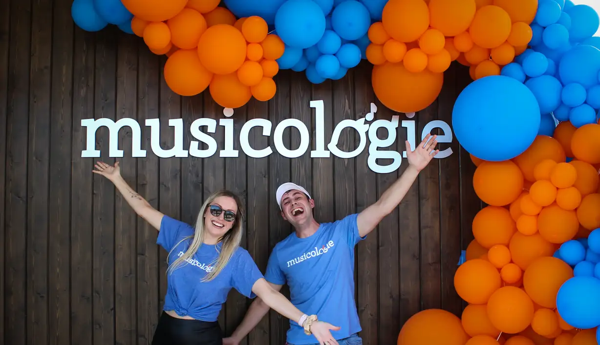 Founder Kay Barker and an excited Community Manager under a brand new Musicologie sign at a grand opening event, surrounded by blue and orange balloons against a modern mood-paneled exterior.
