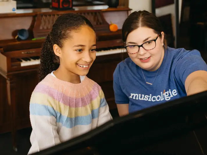 Girl sitting at a piano smiling as her teacher points to a piece of music