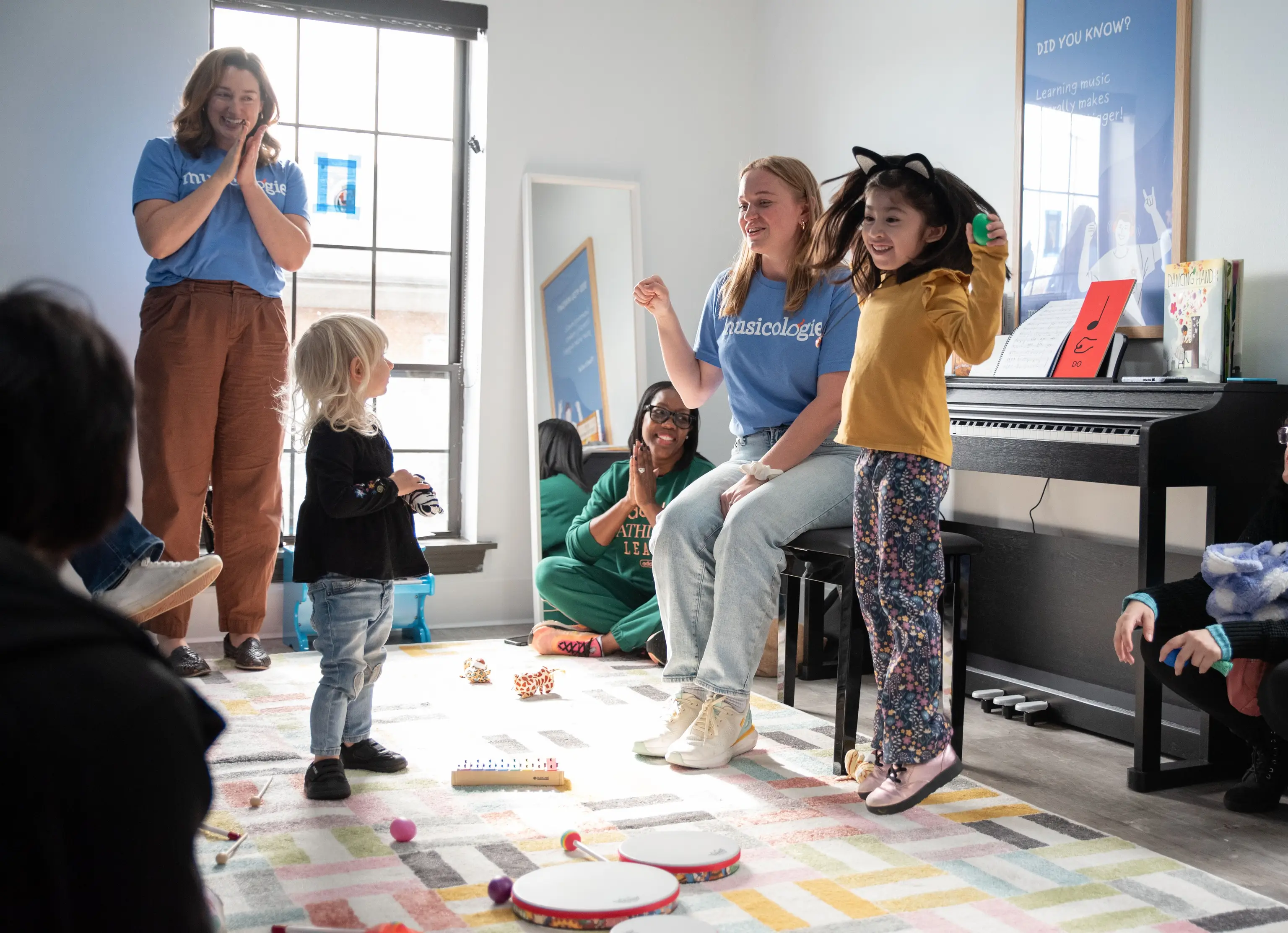 A young girl hops along to music, surrounded by smiling families and teachers at a Musicologie Junior class.
