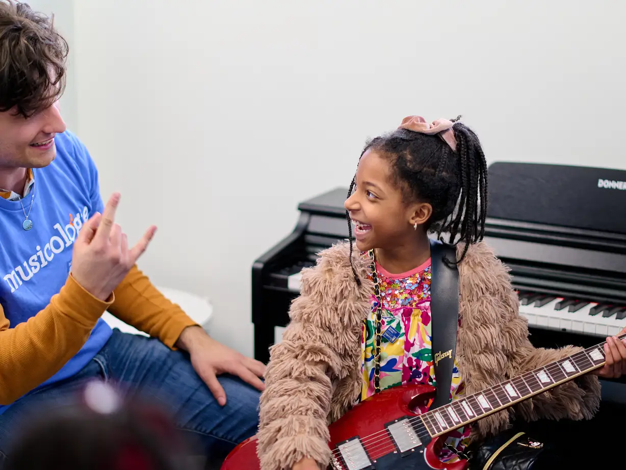 Child holding a red electric guitar and smiling at a teacher at Musicologie.