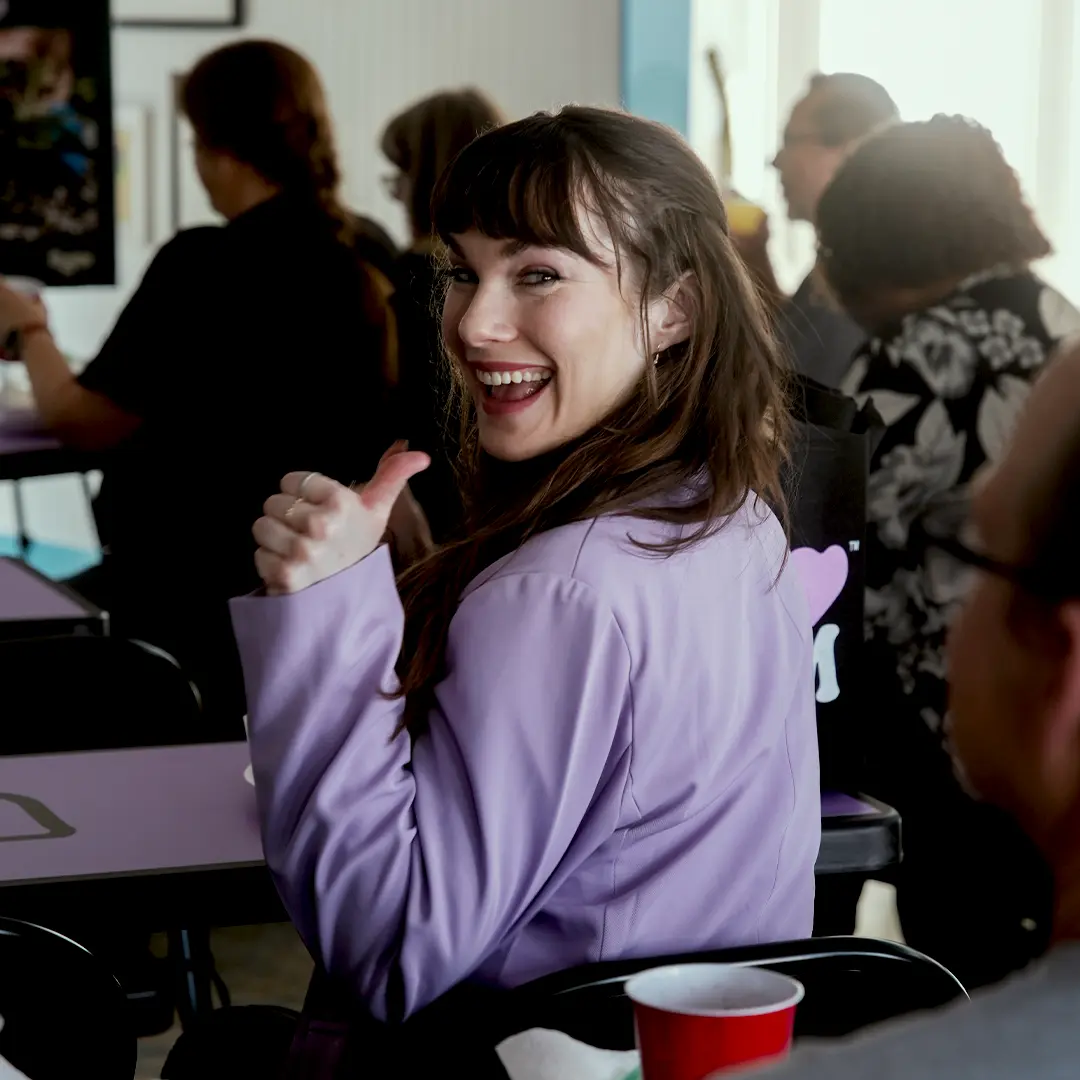 Woman in a purple jacket smiling and giving a thumbs-up while seated at a table in a social setting.