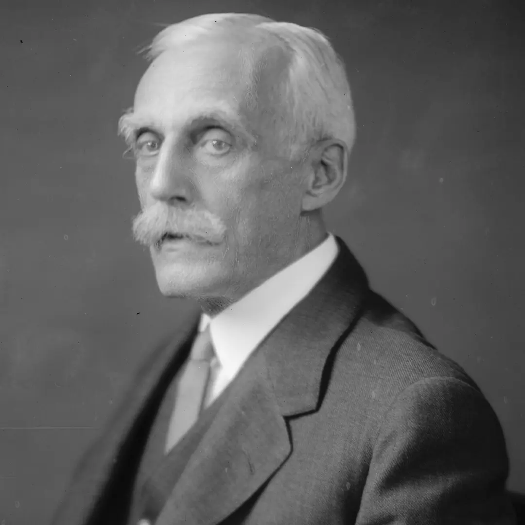 Elderly man with a white mustache and hair, wearing a suit and tie, looking slightly to the side against a plain background.