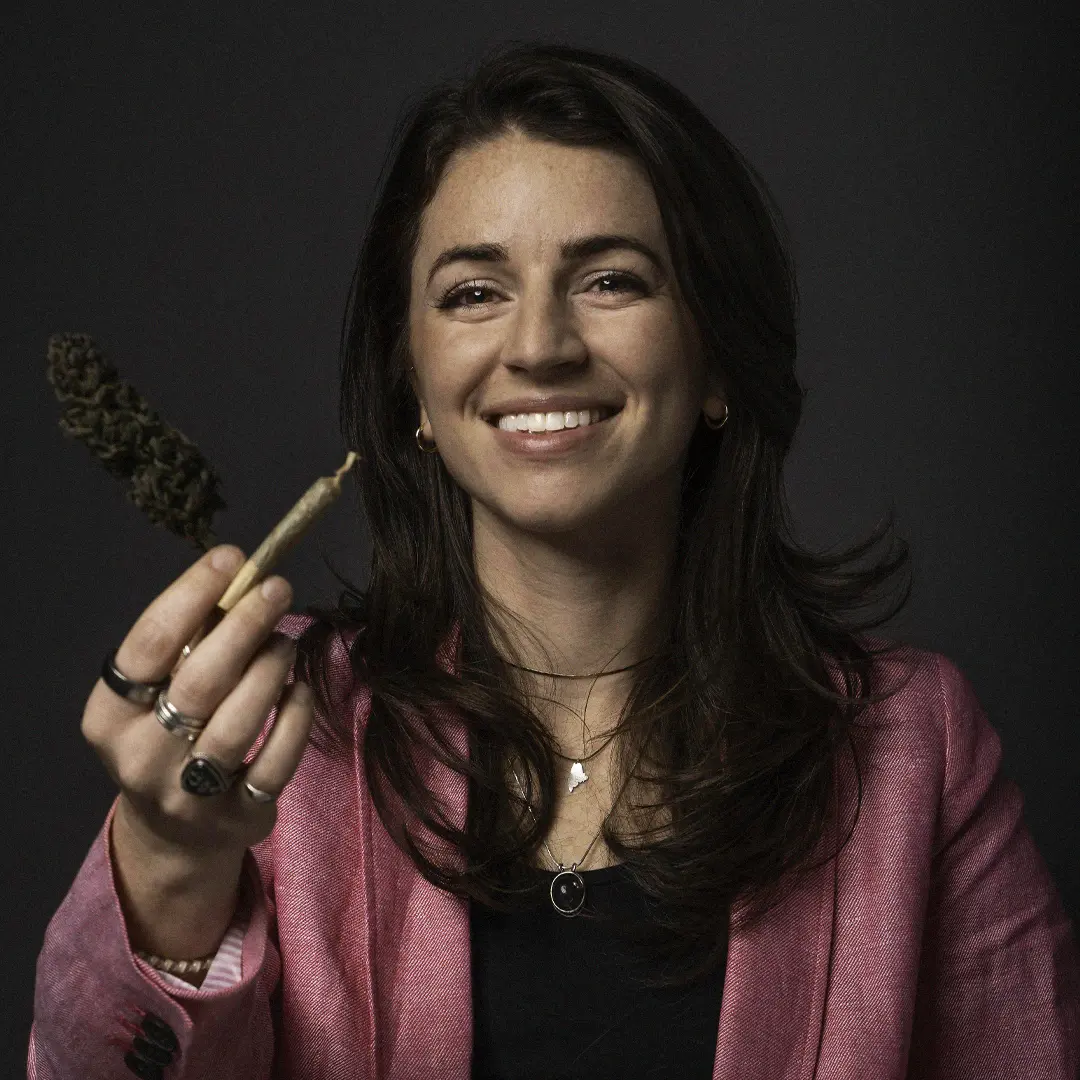 Smiling woman in a pink blazer holding a cannabis joint and bud against a dark background.
