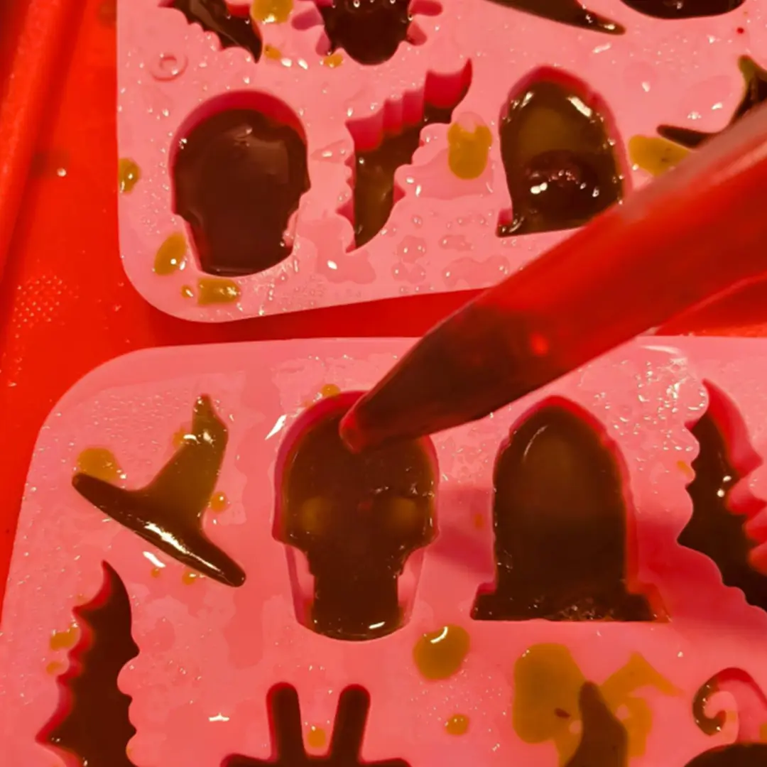 Close-up of a pink silicone mold with Halloween shapes filled with green liquid, including a skull and witch hat. A dropper is adding more liquid.