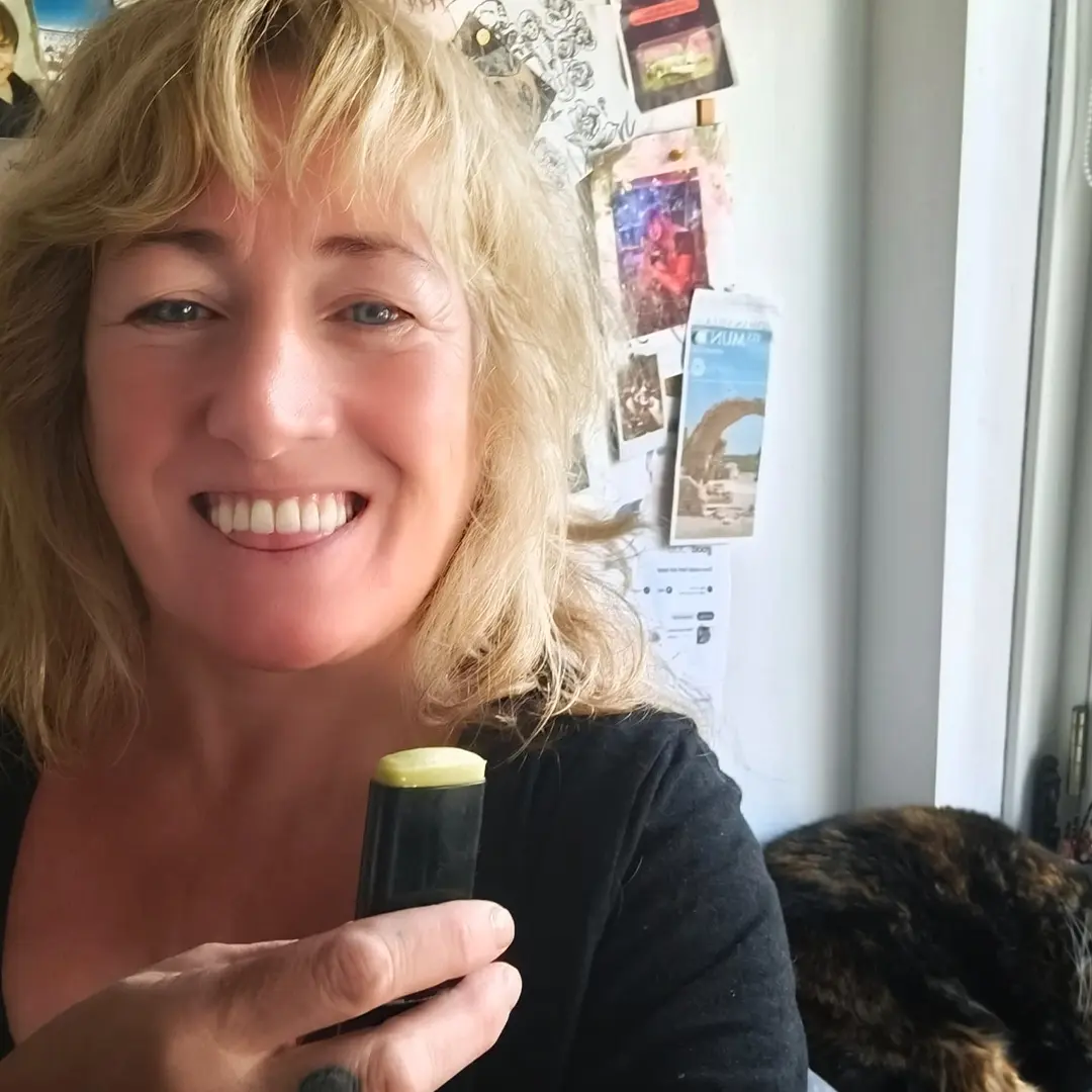 Smiling person holding a small black container with green top, bulletin board behind and a cat curled up by the window.