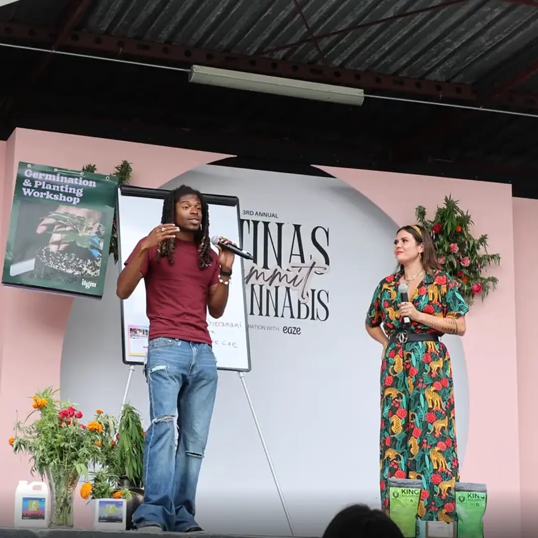 Two people on stage at a cannabis summit, with a presentation board and plants. The man speaks passionately while the woman listens attentively.