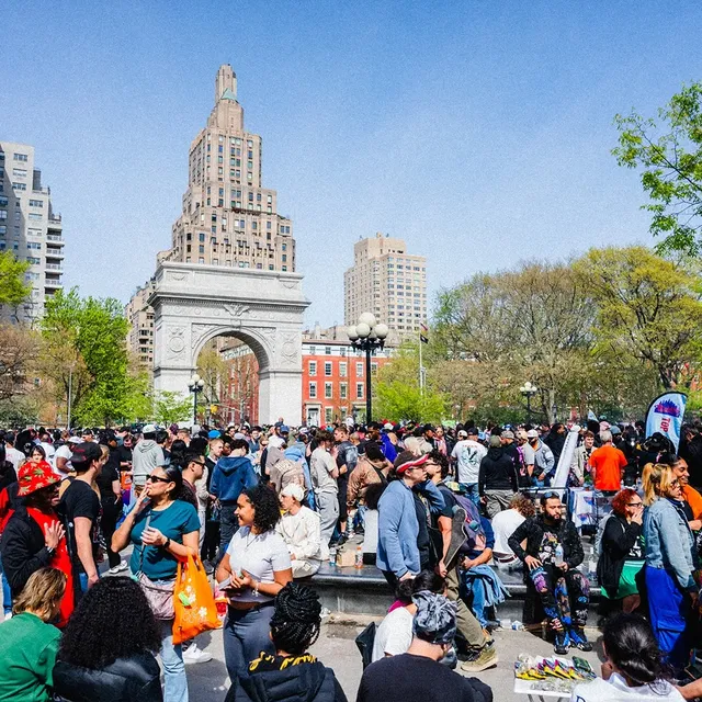 A bustling crowd in a park with a large arch, surrounded by trees and tall buildings under a clear blue sky.