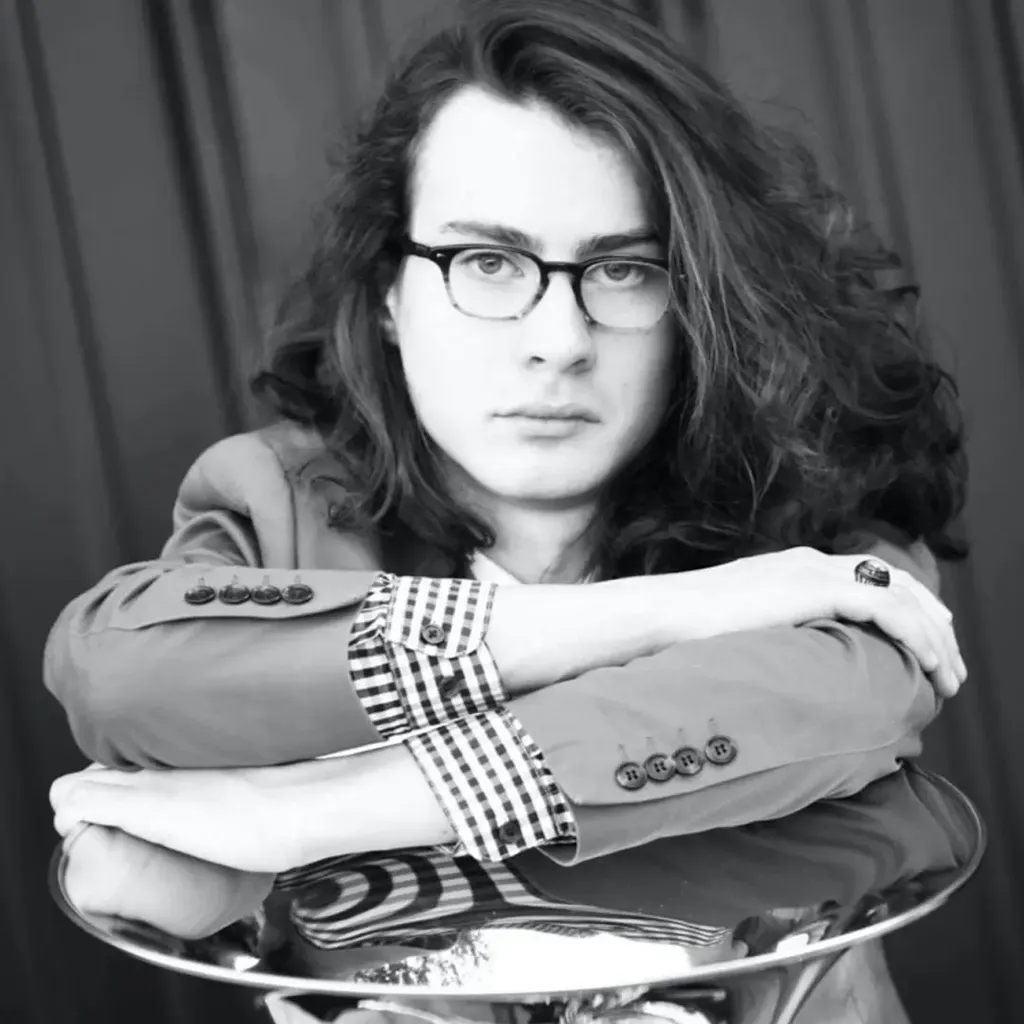 Person with long hair and glasses, wearing a checked shirt and jacket, leans on a reflective surface, looking directly at the camera.