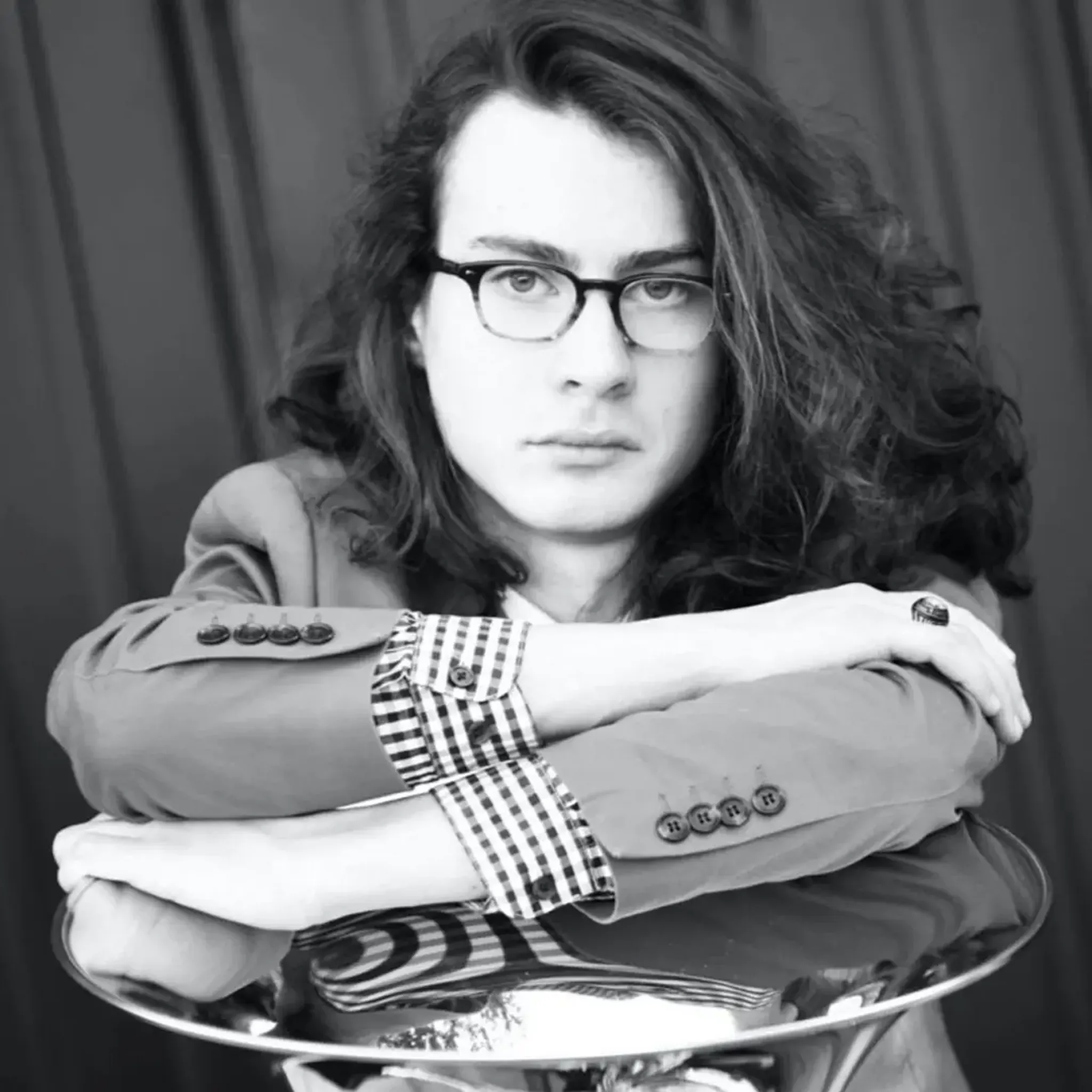 Person with long hair and glasses, wearing a checked shirt and jacket, leans on a reflective surface, looking directly at the camera.