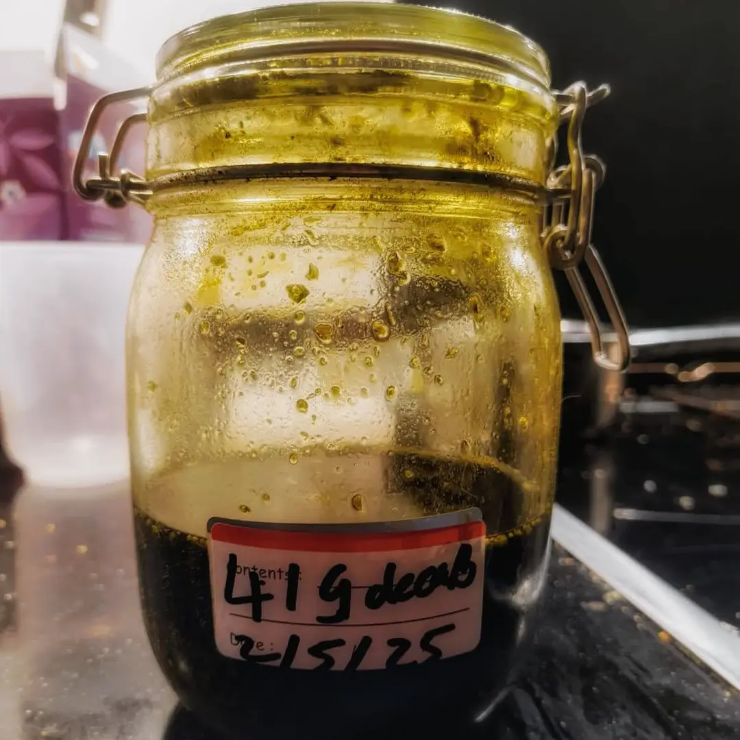 Glass clamp jar containing dark green concentrate with oily residue on the inner walls and a handwritten label.