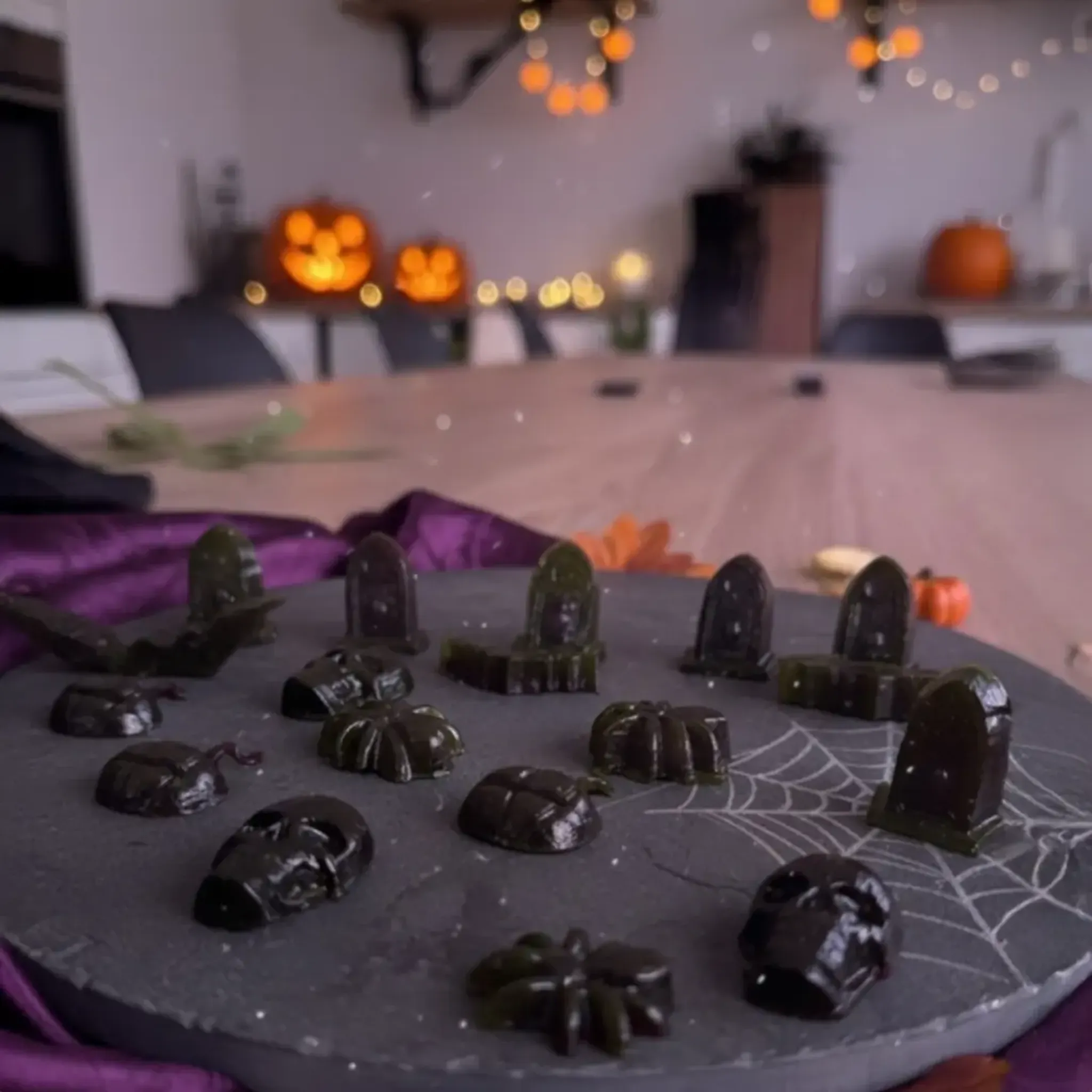 Halloween-themed gummy candies shaped like tombstones, pumpkins, and bats on a plate. Blurred background with pumpkins and string lights.