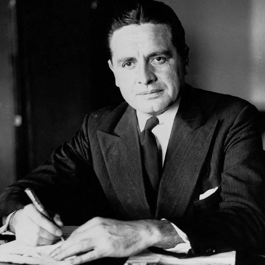A man in a suit and tie sits at a desk, holding a pen and writing on papers, with a focused expression.