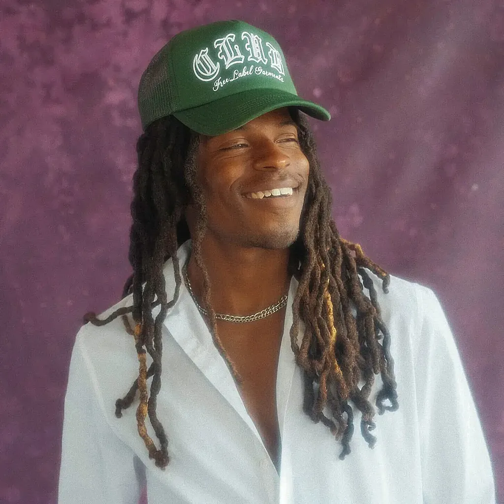Smiling person with long dreadlocks wearing a green cap and white shirt, standing against a purple textured background.