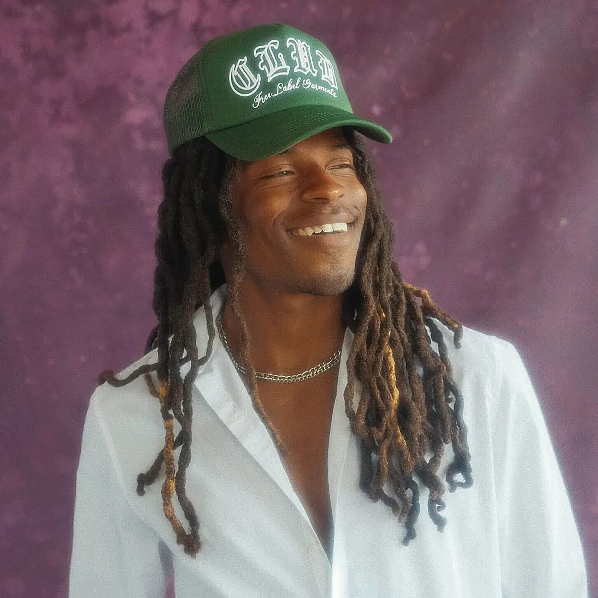 Smiling person with long dreadlocks wearing a green cap and white shirt, standing against a purple textured background.