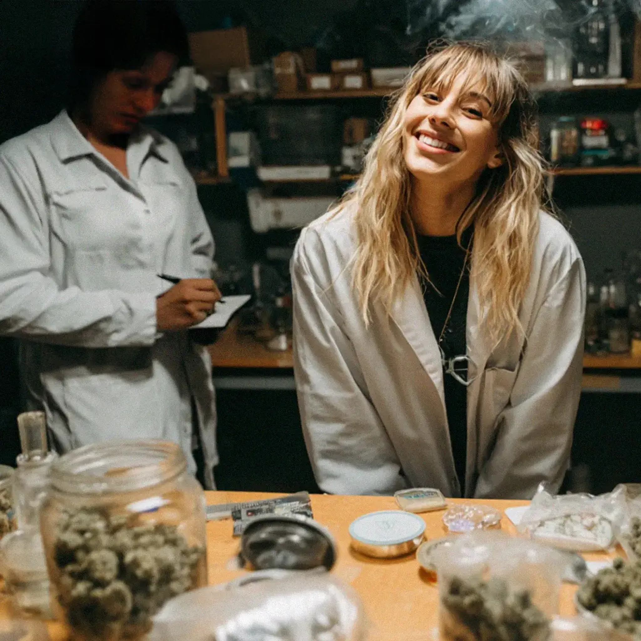 Two people in lab coats in a room with jars of herbs on a table; one is smiling, the other is writing notes.