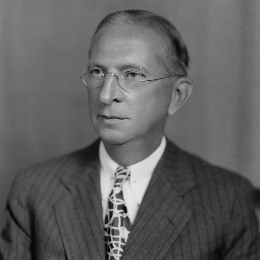 Black and white portrait of an older man in glasses and a suit with a patterned tie, looking to the side against a plain backdrop.