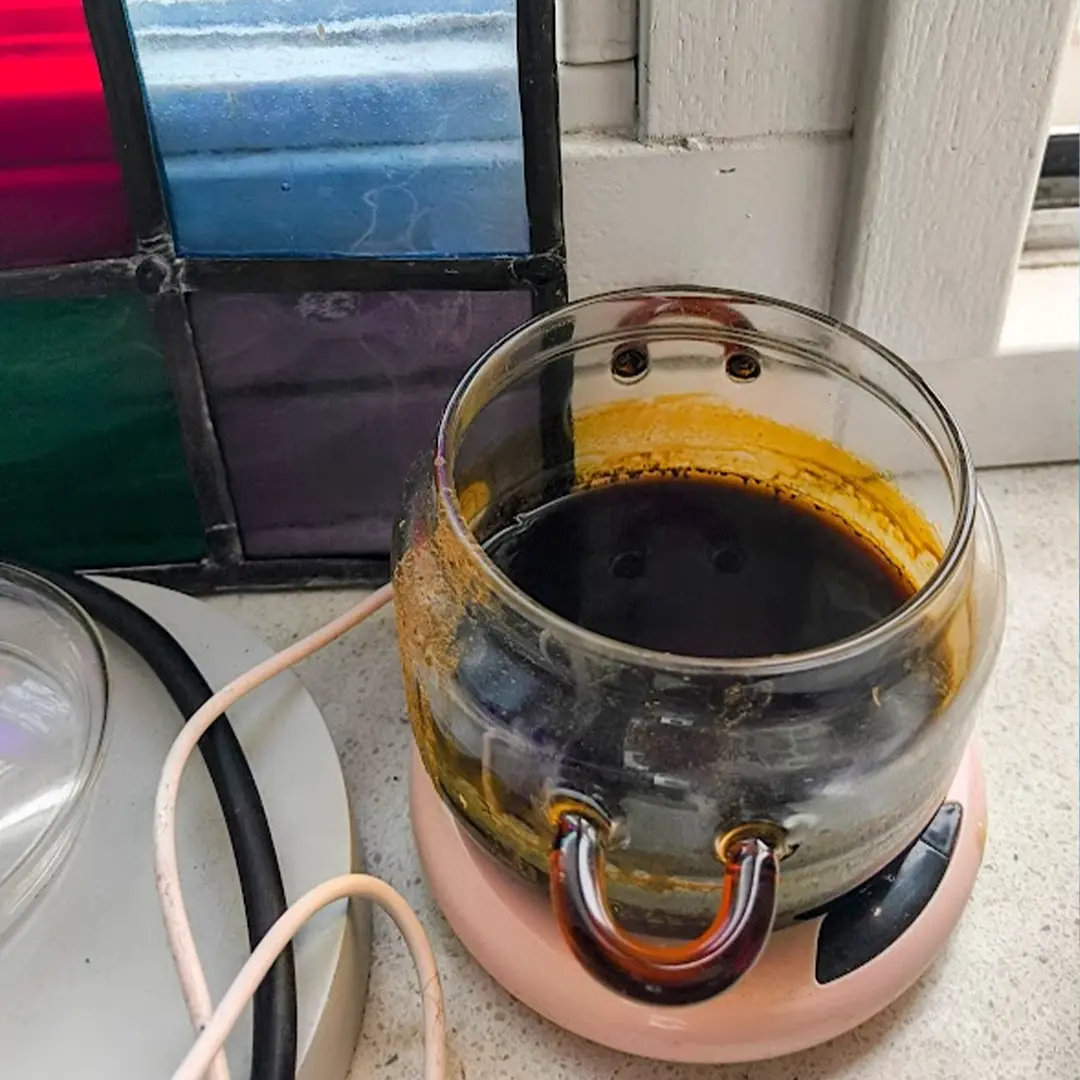 Burnt glass candle jar with dark melted wax and yellow residue sitting on a small pink electric warmer next to stained-glass window and cords.