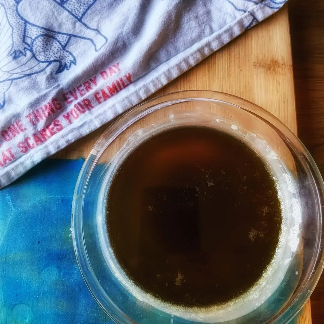 Overhead view of a glass bowl filled with dark liquid on a wooden board, with a blue cloth and white towel with red text at top left.