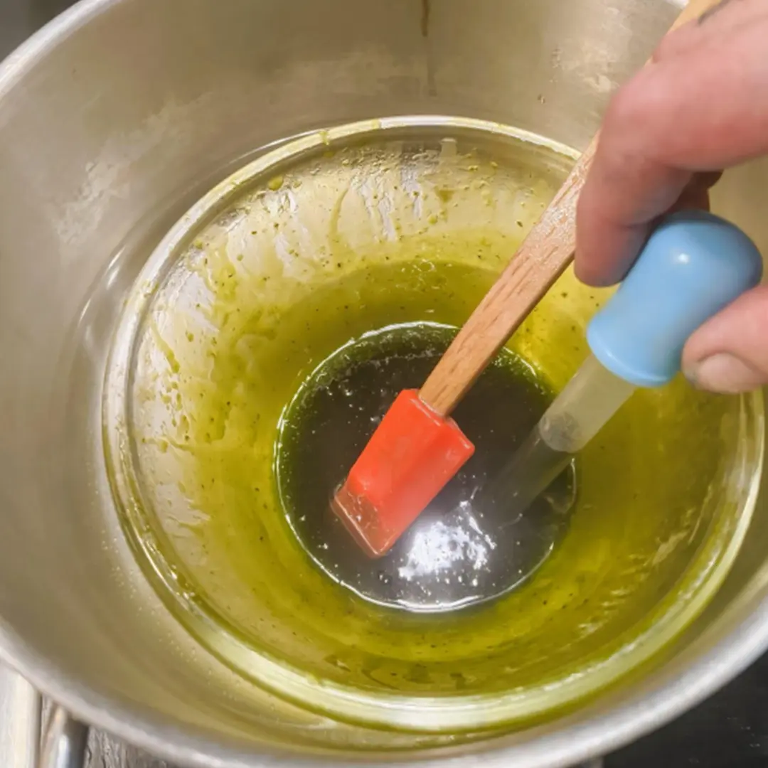 A hand holding a pipette and a spatula over a pot containing green liquid, possibly oil, in a metal container.