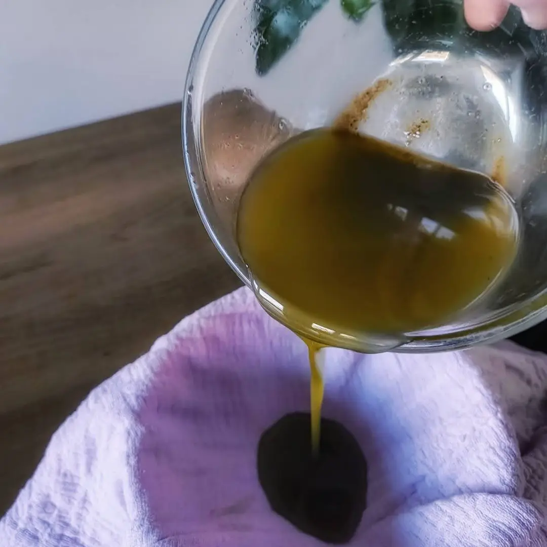 Greenish-brown viscous liquid being poured from a glass bowl onto a purple cloth.