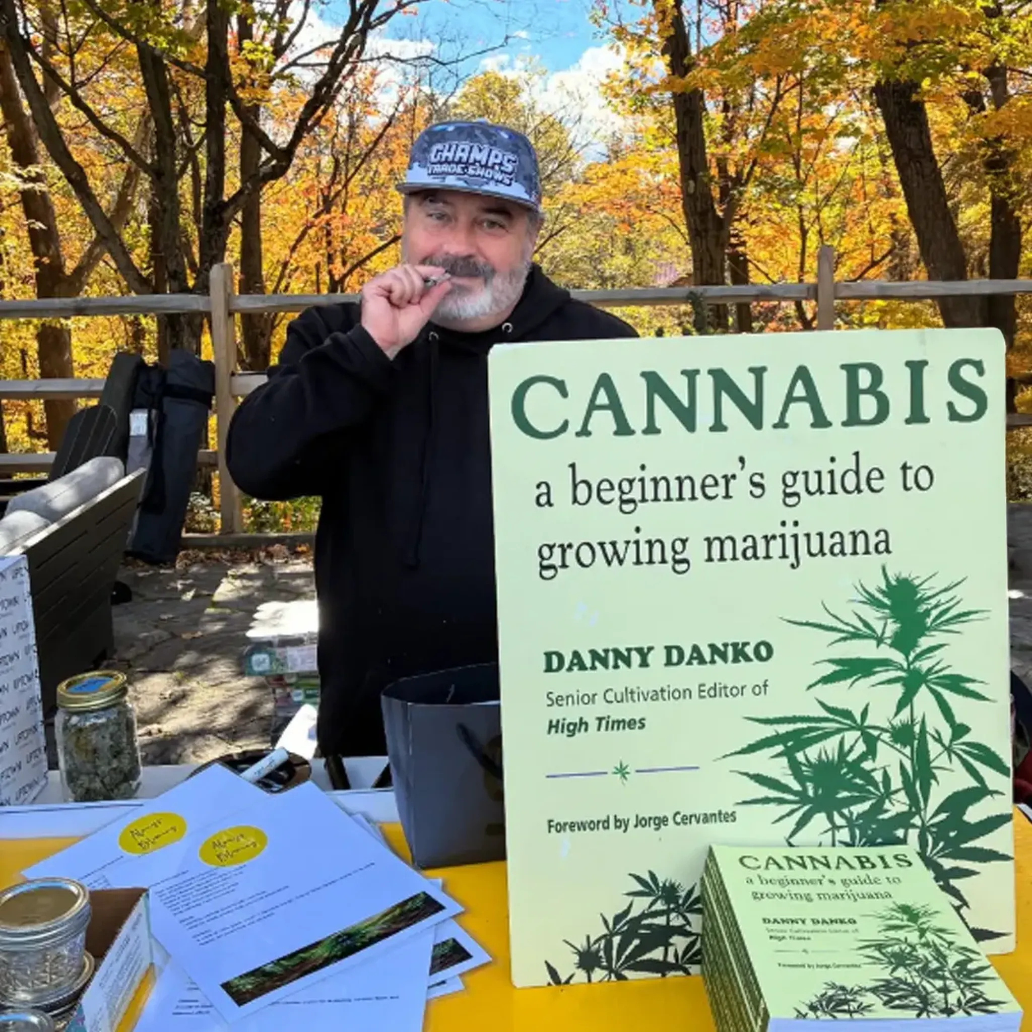 Man at outdoor booth with a cannabis guidebook, wearing a cap, surrounded by autumn trees and promotional materials.