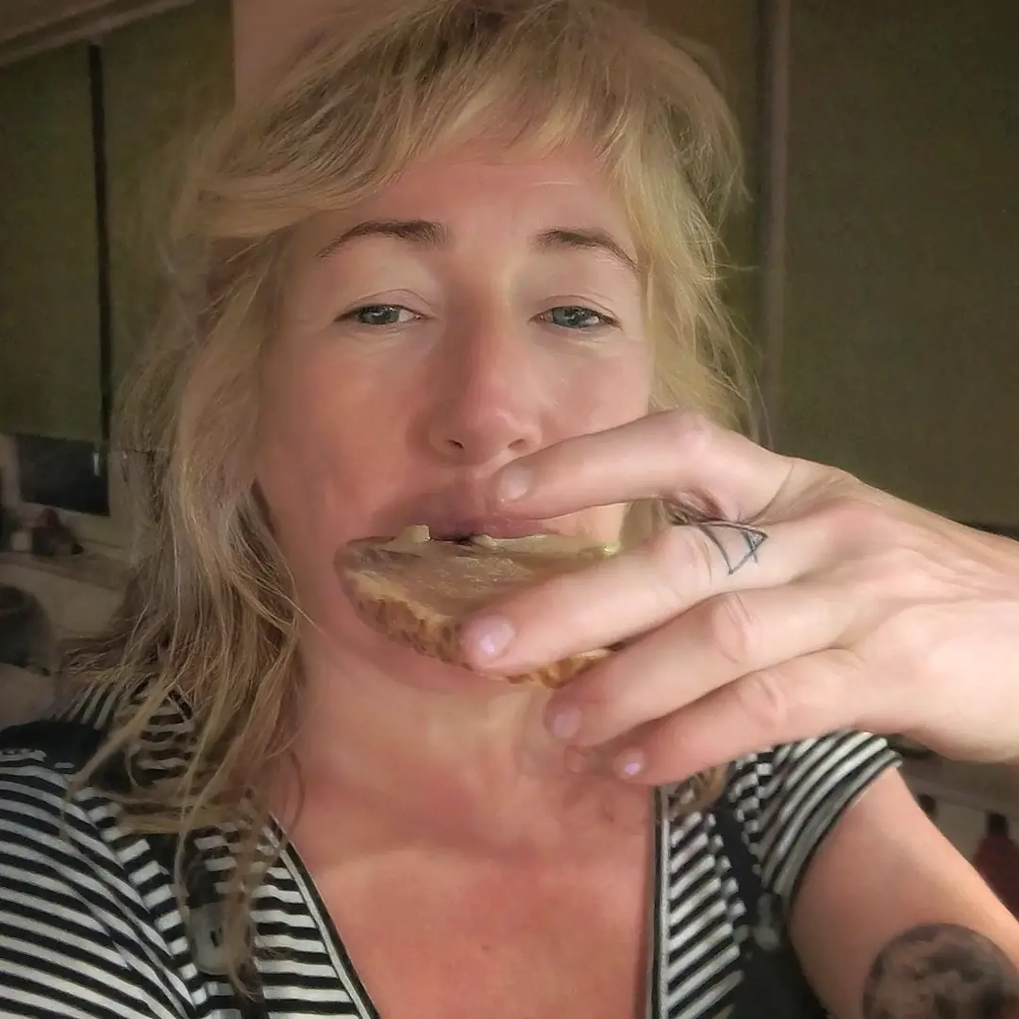 Person with blonde hair eating a slice of bread, wearing a striped shirt, with a tattoo on their finger, in an indoor setting.