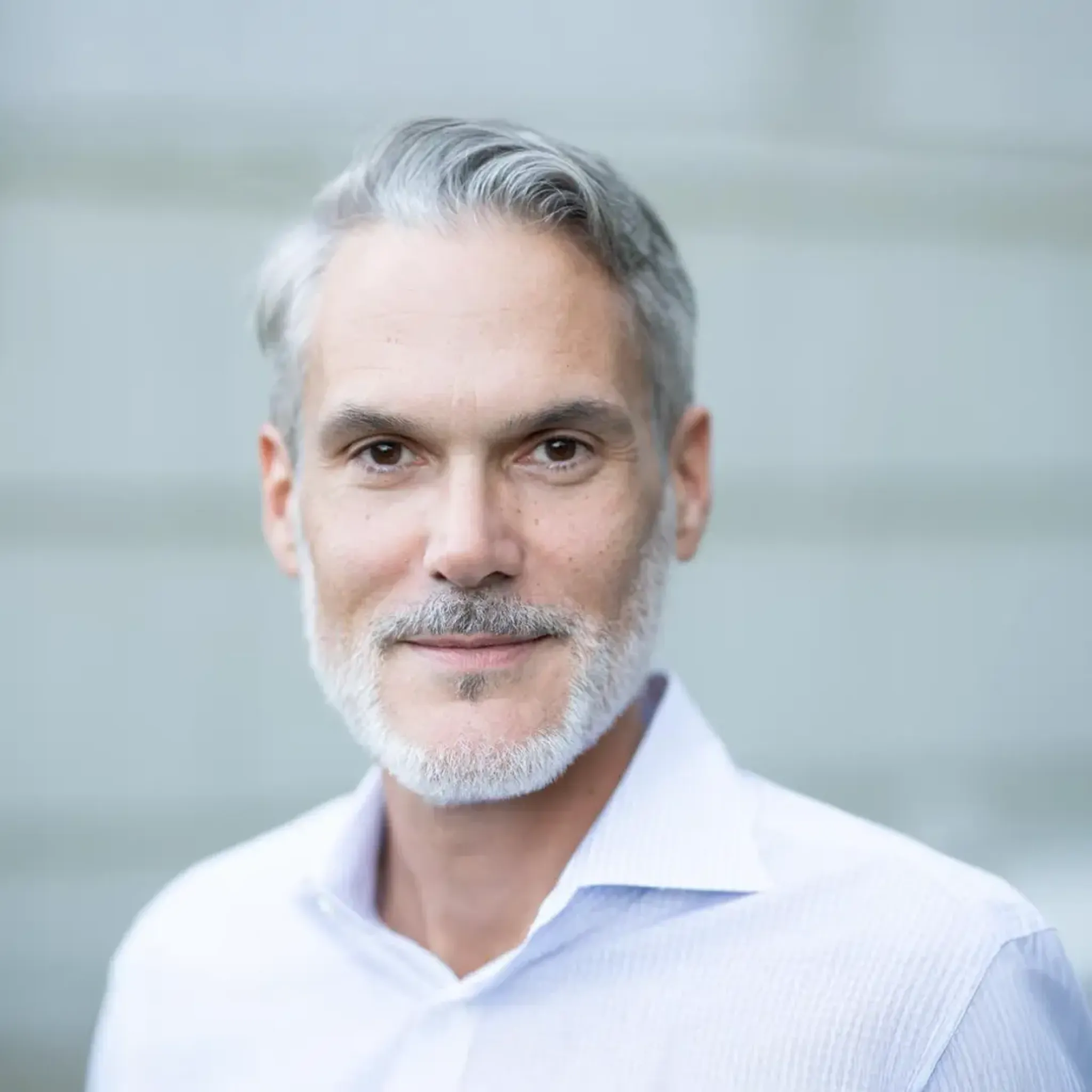 Professional headshot of a smiling middle-aged man with gray hair and beard wearing a light dress shirt.