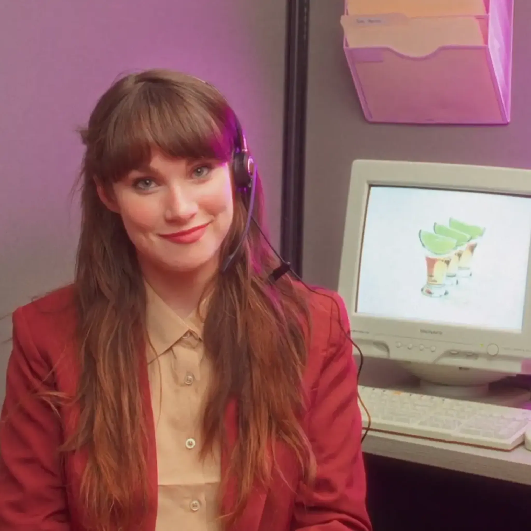 Woman in a red blazer with a headset, sitting at a desk with a vintage computer displaying a cocktail image. Office setting with soft lighting.