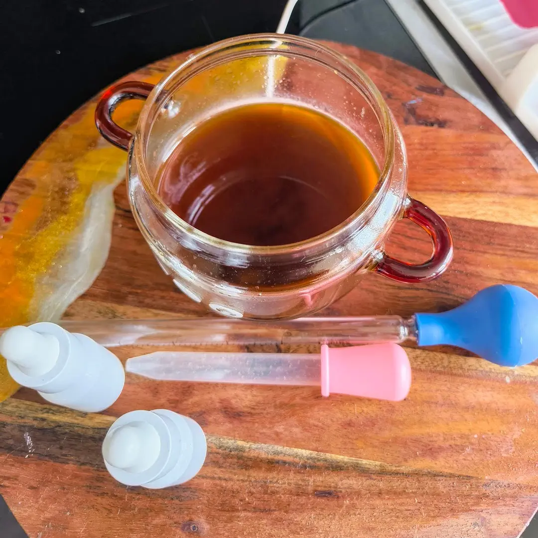 Top-down view of a glass cup with dark liquid on a wooden board, two pipettes with pink and blue bulbs and white caps.