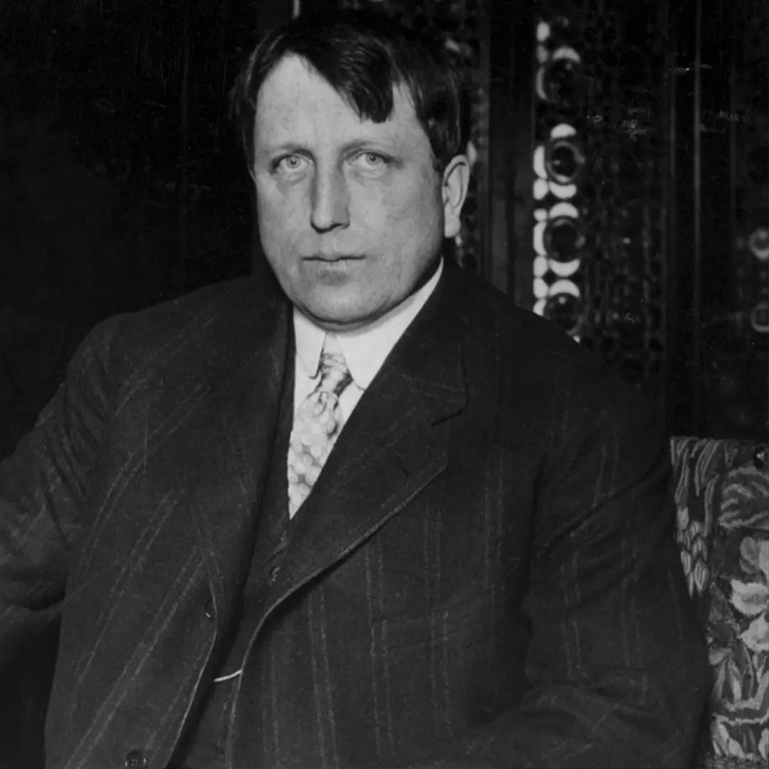 A man in a suit and tie sits in a chair, looking directly at the camera with a neutral expression. The background features ornate patterns.