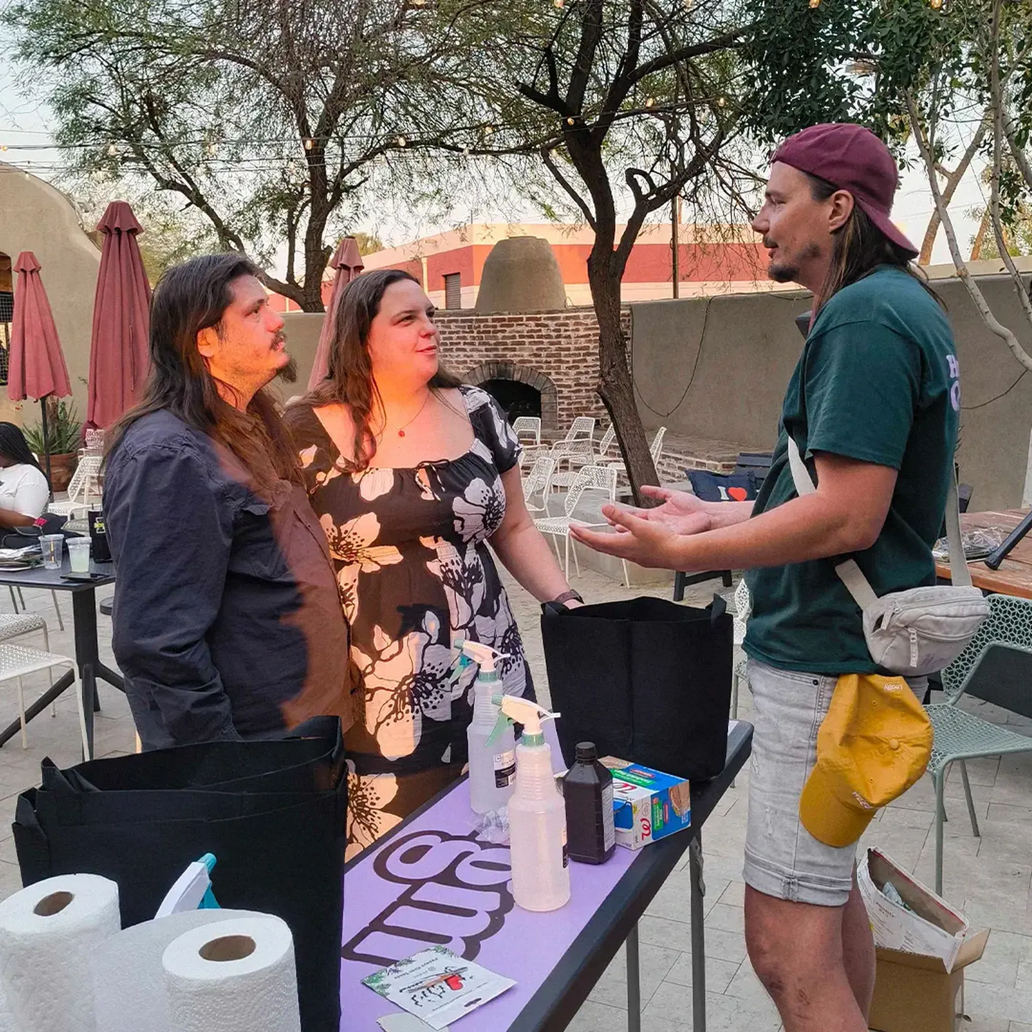Three people converse outdoors near tables with supplies like spray bottles and paper towels. Trees and umbrellas are in the background.